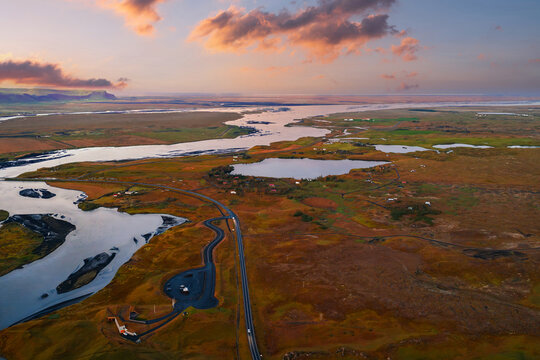 Aerial view of Icelandic wetlands near Vik at sunset, with braided rivers, lagoons, oxbow channels, a road through golden mossy plains, and sparse farms under warm light.