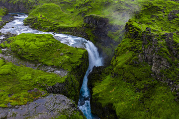 Aerial view shows a turquoise river dropping into a basalt gorge in Iceland highlands near Haifoss or Sigoldugljufur, with mist, mossy lava, and overcast light.