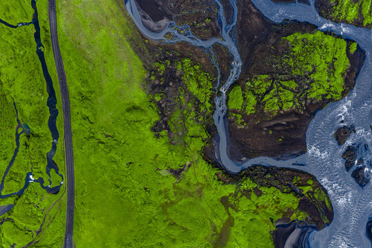 Aerial view of neon green moss on dark lava with braided streams near Vik, Iceland. A narrow road lines the left, rivers merge right in crisp daylight contrast.
