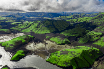 Aerial view of Landmannalaugar in Iceland shows mossy rhyolite hills, volcanic cones, dark lava fields, and braided rivers under broken clouds in daylight wide angle