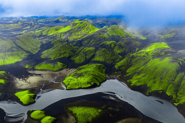 Fototapeta premium Aerial view of Landmannalaugar, Iceland, shows neon green moss on volcanic ridges and craters, black lava sands, and a braided glacial river under low clouds and diffuse light.