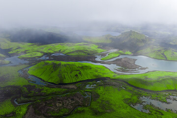 Aerial view of moss green hills, braided rivers, lava fields, and rhyolite ridges in Iceland Highlands near Landmannalaugar, with low clouds and diffused daylight.