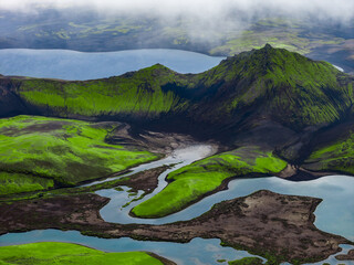 Aerial view of Iceland highlands shows moss green ridges, black lava, crater rims, and braided lakes in Landmannalaugar or Fjallabak under low cloud with soft light.