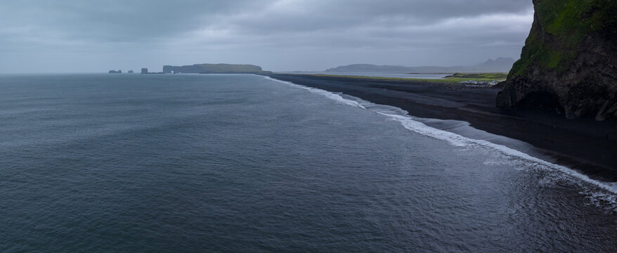 Overcast aerial view of Reynisfjara near Vik i Myrdal, Iceland. Gentle waves trace white lines on black sand, Reynisdrangar rise left, mossy cliffs frame right.
