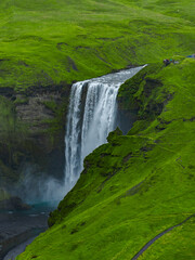 Fototapeta premium A broad Skogafoss waterfall drops over a rocky cliff into a misty gorge in southern Iceland. Mossy hills, winding footpaths, and tiny hikers show scale in soft overcast light.