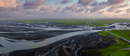 Aerial view shows braided glacial river across dark volcanic sands and green moss, a straight bridge and highway on Iceland south coast sandur at pastel dusk.