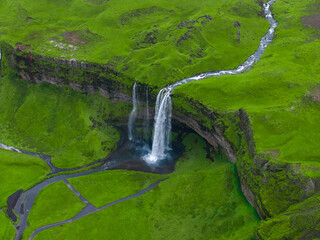 Aerial view shows Seljalandsfoss in southern Iceland as white water drops before a shallow cave into a dark pool, with a thin river on the plateau under soft overcast light.