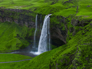 Fototapeta premium Aerial view shows Seljalandsfoss on Iceland South Coast, white water drops over mossy basalt into a shallow pool, footpath and recess behind the falls are visible in soft light.