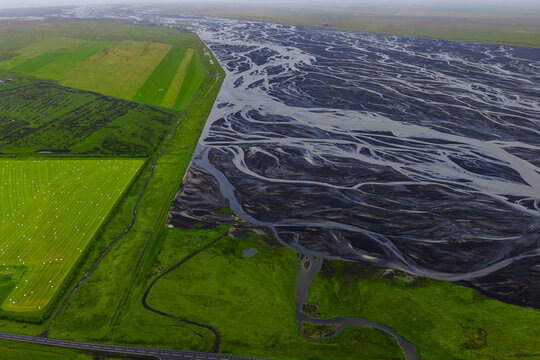 Aerial view shows green fields with hay bales and levees beside a black braided river plain on Iceland south coast near Skeidararsandur, with soft diffused light.