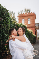 Naklejka premium Wedding photography, portrait of happy smiling newlyweds, a young groom and a beautiful sweet brunette bride, tenderly hugging outdoors on a street in the city, in a park.