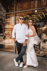 Fototapeta premium Wedding photography, portrait of happy cool newlyweds in sunglasses, young stylish groom and beautiful bride, standing against glass background of store on street in city.