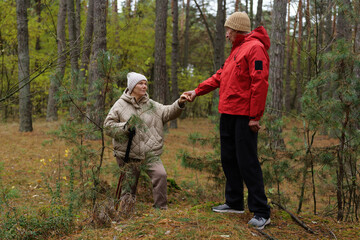 Fototapeta premium An elderly woman sits among green vegetation while a man in a red jacket holds her hand. They enjoy the peaceful atmosphere of the forest