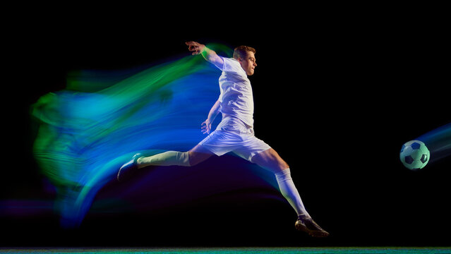 Soccer player kicking ball with green and blue neon motion trail on dark field. Concept of performance, ambition, and athletic energy in sports campaigns and digital design visuals.