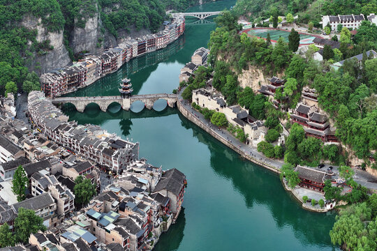 Aerial view of the ancient town's weathered buildings and arched bridges reflected in the jade river winding through verdant hills, Zhenyuan, Guizhou, China.