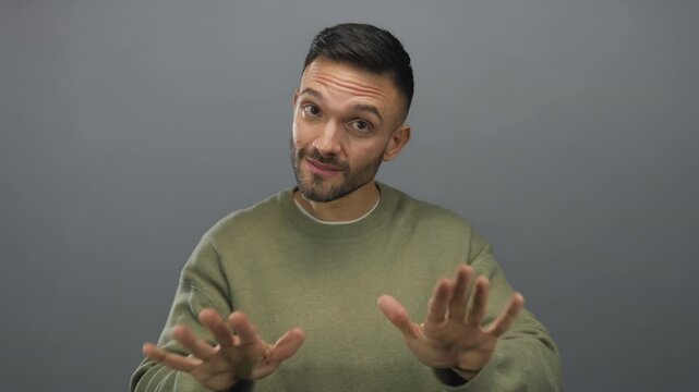 Hispanic man gesturing calm with hands against a plain grey background, wearing casual green sweater, expressing tranquility and reassurance in a neutral setting.