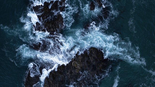 Aerial view of dark rocks embraced by foamy, turquoise waves, a dramatic clash of textures and tones where the ocean meets the rugged coast, California, United States.