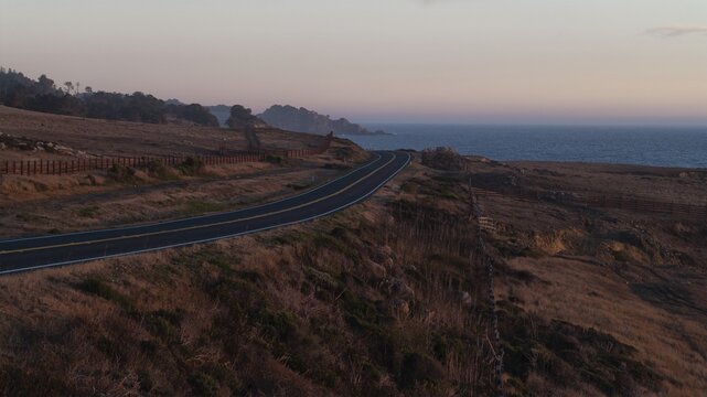 Aerial view of a winding coastal road cutting through the golden grasslands, as the pacific ocean kisses the horizon near the Mendocino Headlands State Park, Mendocino, California, United States.