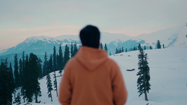 4K shot of a defocused Indian male tourist looking at the snowy Himalayan mountain peaks during the sunset in winter season at Gulmarg in Jammu and Kashmir, India. Tourist enjoying scenic views.	