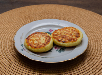 Two golden brown cheesecakes or cottage cheese pancakes on a floral plate. Homemade breakfast or dessert served on a rustic woven placemat.