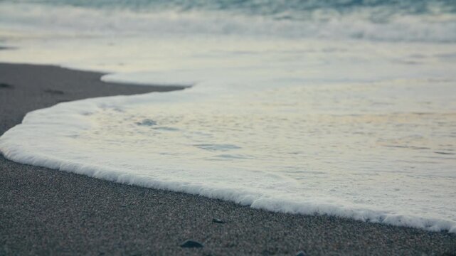 Close-up of a gentle wave washing away footprints on the beach, symbolizing impermanence and calm, captured in soft evening light.