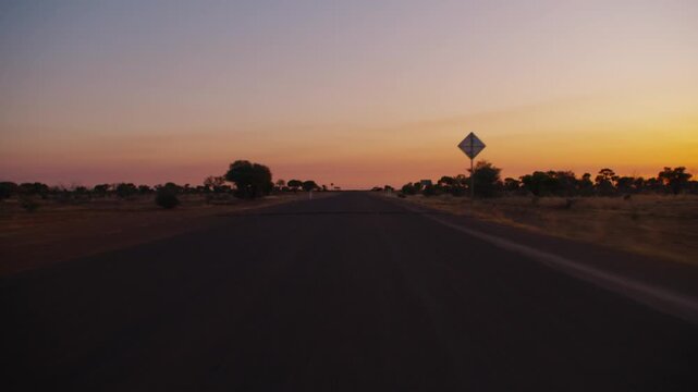 The iconic Australian outback sunset with a country road and the silhouettes of large trees as a car drives down the long stretch of highway.