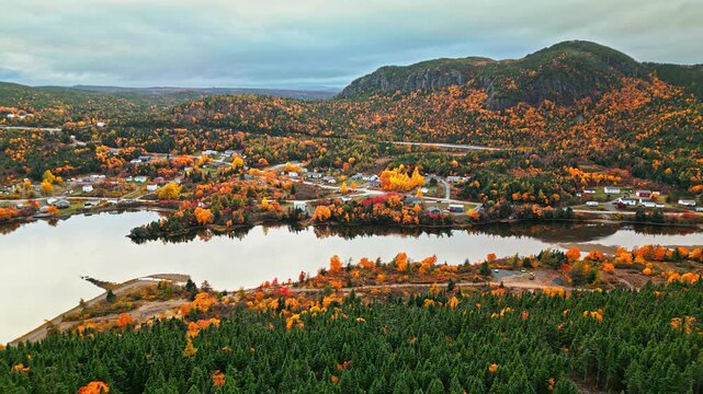 Drone aerial sweeps over the Newfoundland town of North West Brook, showing houses, roads and a lake shimmering with autumn colours beneath a rugged mountain.