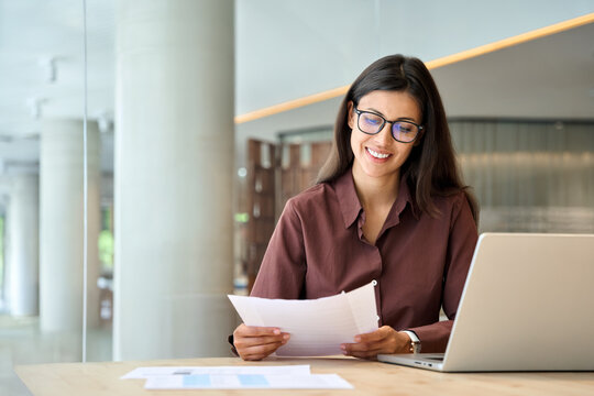 Smiling latin hispanic business woman working on laptop computer reading financial document report in office. Accountant entrepreneur manager businesswoman doing paperwork using pc. Copy space