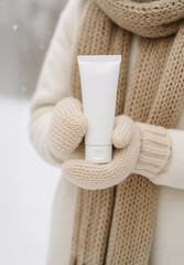 Close-up of woman in warm beige mittens holding a white tube of hand cream outdoors in winter. Skincare and protection concept for cold weather, moisturizing and beauty product mockup.
