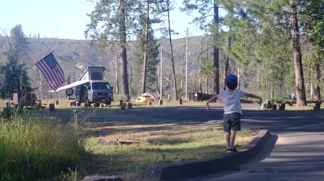Young boy balancing on curb at a summer campsite in a national park, a microbus camper van with an american flag under a sunny sky, enjoying outdoor leisure and family vacation