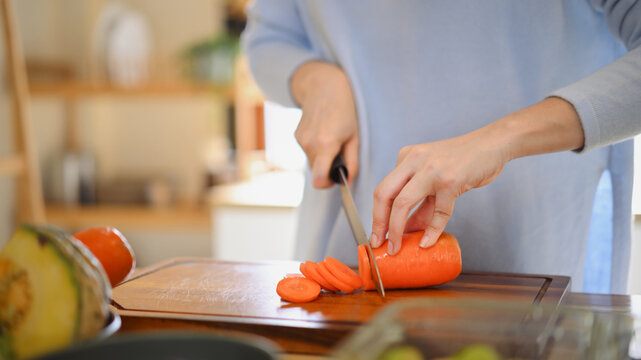 Closeup of young female hands chopping fresh orange carrot on board while in modern kitchen