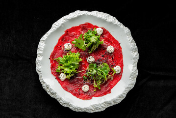 Top-down view of beef carpaccio, a classic Italian appetizer, with thin slices of raw meat, garnished with arugula, cream cheese, and spices on a white plate against a black background