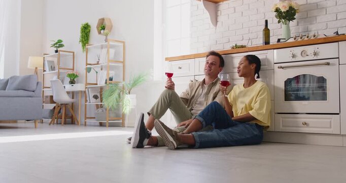 Happy couple toasting and clinking red wine glasses together, cheerful two people talking at home. Young man and woman sitting on kitchen floor, sharing drink and conversation, enjoying romance