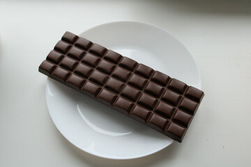 Dark chocolate bar with rectangular segments placed diagonally on white plate showing classic chocolate bar design and texture.