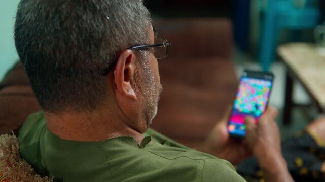 A side profile close-up of a middle-aged Asian man with a beard and glasses, sitting on a sofa and intently playing a mobile game on his smartphone.