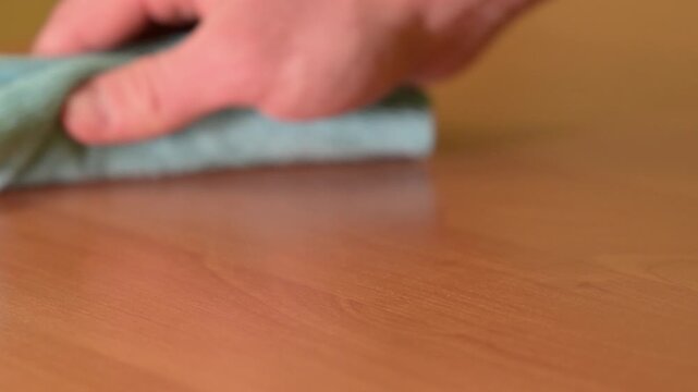 Hand wiping a wooden table surface. Close-up of a man's hand pulling a green microfibre cloth across a laminated brown table surface, leaving a thick layer of dirt and dust on the fabric