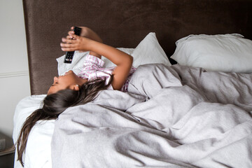 A young girl is laying in bed with a remote in her mouth