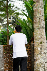 Man wearing a plain white t-shirt standing outdoors near a concrete fence and tropical plants, showing minimal and natural lifestyle fashion concept.