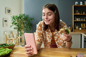 Portrait of teenage girl smiling while holding sandwich and looking at smartphone in kitchen, showing cheerful expression and engaging with device during meal
