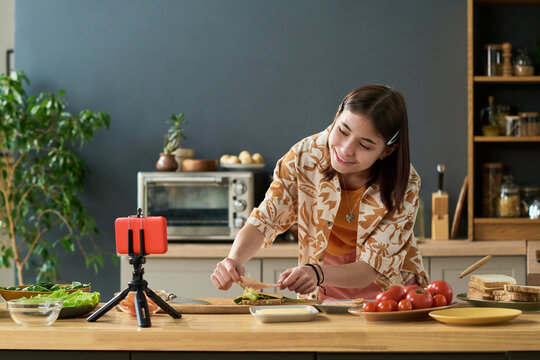 Teenage girl smiling while preparing vegetables on kitchen counter, recording cooking video with smartphone on tripod, looking down at cutting board, modern home kitchen background