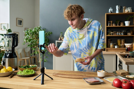 Teenage boy preparing sandwich while recording video on smartphone in modern kitchen, holding bread and interacting with device, fresh vegetables and ingredients on counter