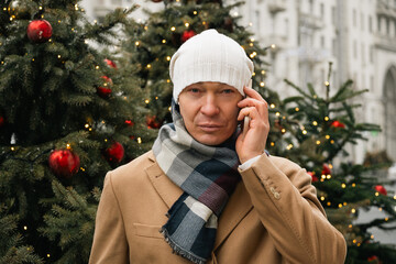 Adult handsome man stands thoughtfully with phone and call on street near Christmas trees in winter attire in modern city. Xmas holiday.