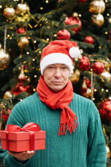Happy man wearing green sweater holding red gift and stand near Christmas tree. Xmas Holiday. Close up.