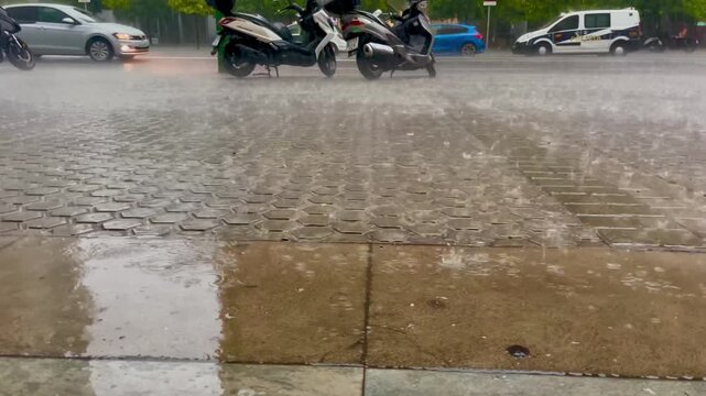 Raindrops falling into puddles on the sidewalk near the road with passing cars. Moody urban rain scene.
