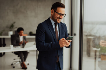 A man in an office typing on his phone near a window, while his female colleague sits at a desk behind him

