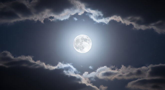 A brilliant full moon glowing intensely in the center of the night sky, framed by dramatic, silhouetted clouds in a celestial display