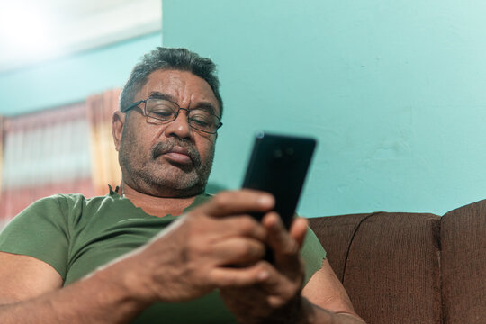 A side profile close-up of a middle-aged Asian man with a beard and glasses, sitting on a sofa and intently playing a mobile game on his smartphone.
