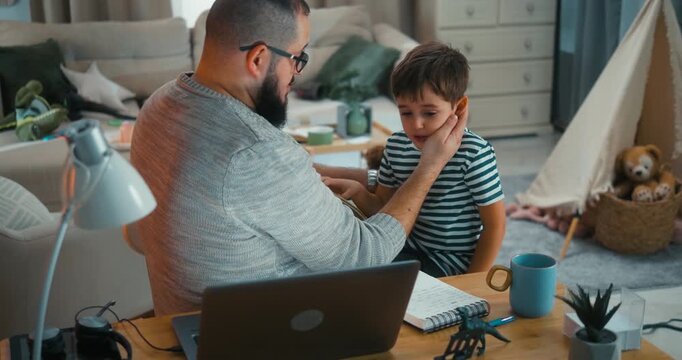 Father successfully comforts his child after a minor upset and returns to work at his desk. Real remote work and parenting, productivity and balancing family life in a warm everyday home scene.