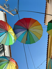 Upward view of colorful umbrellas suspended above a narrow street under a bright blue sky.