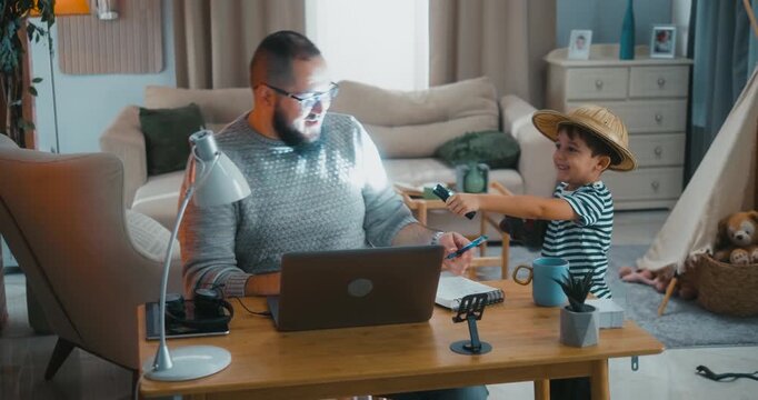 Playful boy interrupts his father working at the home desk, turning the moment into a fun game. Real everyday balance of remote work, parenthood and positive family interaction.