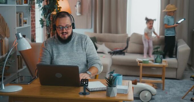 Father on a business video conference call from home while kids jump, make noise and disrupt behind him. Real work-from-home struggle, multitasking and chaotic parenting in daily life.